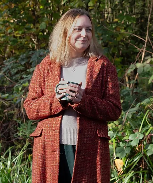 Natalie Cooper holding a mug, she's standing in a long orange winter coat outside with a hedge behind her.