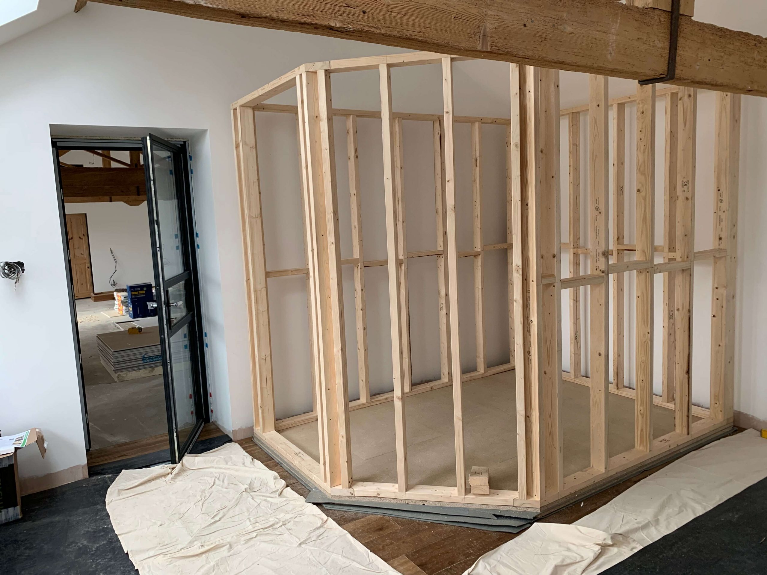 Wooden framework for a home voiceover recording booth resting on a double layer of TecSound acoustic membrane, positioned in the corner of a room next to an open glass door, with a large structural wooden beam in the foreground.