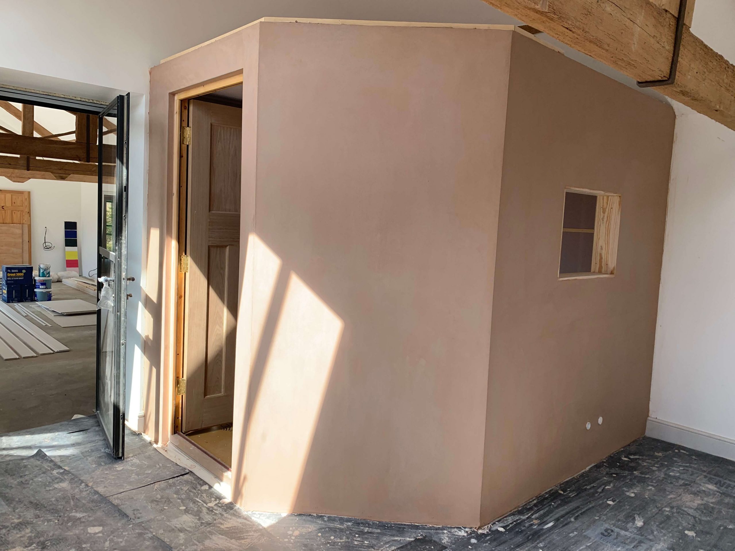 Fully formed home voiceover recording booth with an outer layer of plaster, oak door open into the booth, window frame in place but glass not yet installed.
