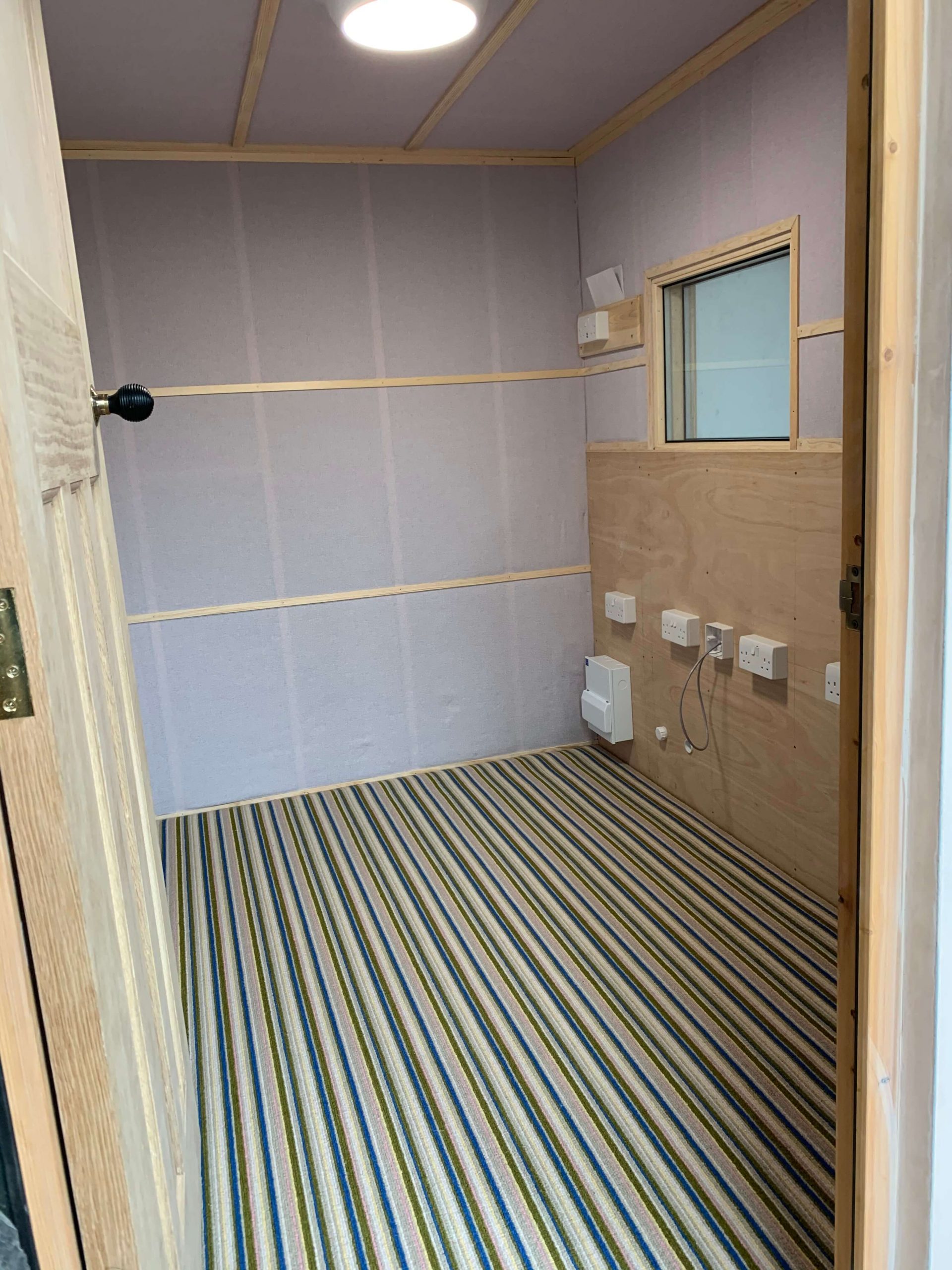 Completed home voiceover recording booth interior viewed through the open oak door, showing pale pink acoustic fabric covering the walls and ceiling, carpet on the floor, installed window glass, and a small consumer unit with electrics in place.