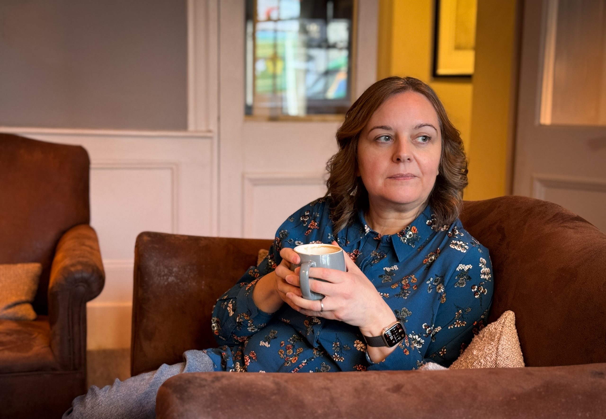 Natalie, a middle aged woman with brown hair wearing a blue patterned shirt, sitting on a brown leather sofa drinking a coffee and loking thoughtful